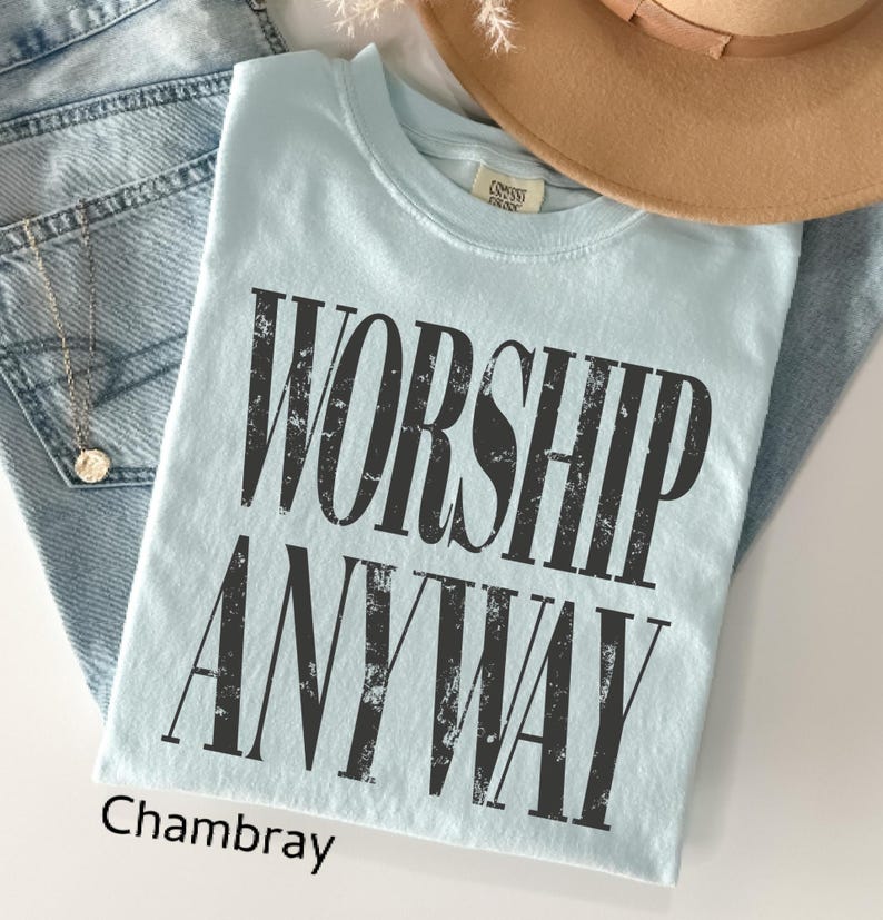 May include: A light blue t-shirt with the words "WORSHIP ANYWAY" in distressed black lettering. The shirt is folded and rests on a pair of blue jeans. A tan hat is visible in the background.