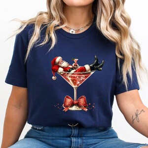 May include: A navy blue t-shirt with a graphic of Santa Claus relaxing in a martini glass with a red bow.