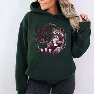 May include: A dark green hoodie with a graphic of a raccoon holding a cup of coffee. The text "Cute But Feral" is printed on the hoodie.