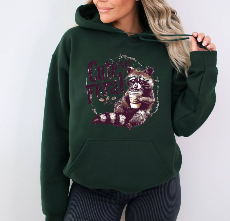 May include: A dark green hoodie with a graphic of a raccoon holding a cup of coffee. The text "Cute But Feral" is printed on the hoodie.