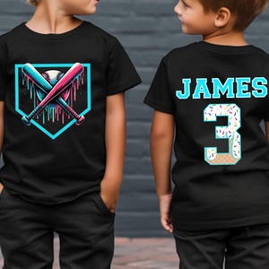 May include: Two black t-shirts with a baseball theme. One shirt features a baseball bat and ball with dripping paint, the other shirt has the name "JAMES" and the number "3" with sprinkles.