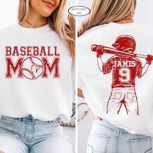 May include: White sweatshirt with red baseball-themed graphics. The front features "BASEBALL MOM" with a baseball and heart. The back displays a baseball player with the name "JAMES" and the number "9".