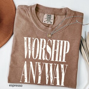 May include: Espresso-colored t-shirt with the words "WORSHIP ANYWAY" in large white letters. The shirt has a crew neck and a small tag at the neckline. A silver necklace with a pendant rests on the shirt. The shirt is folded.