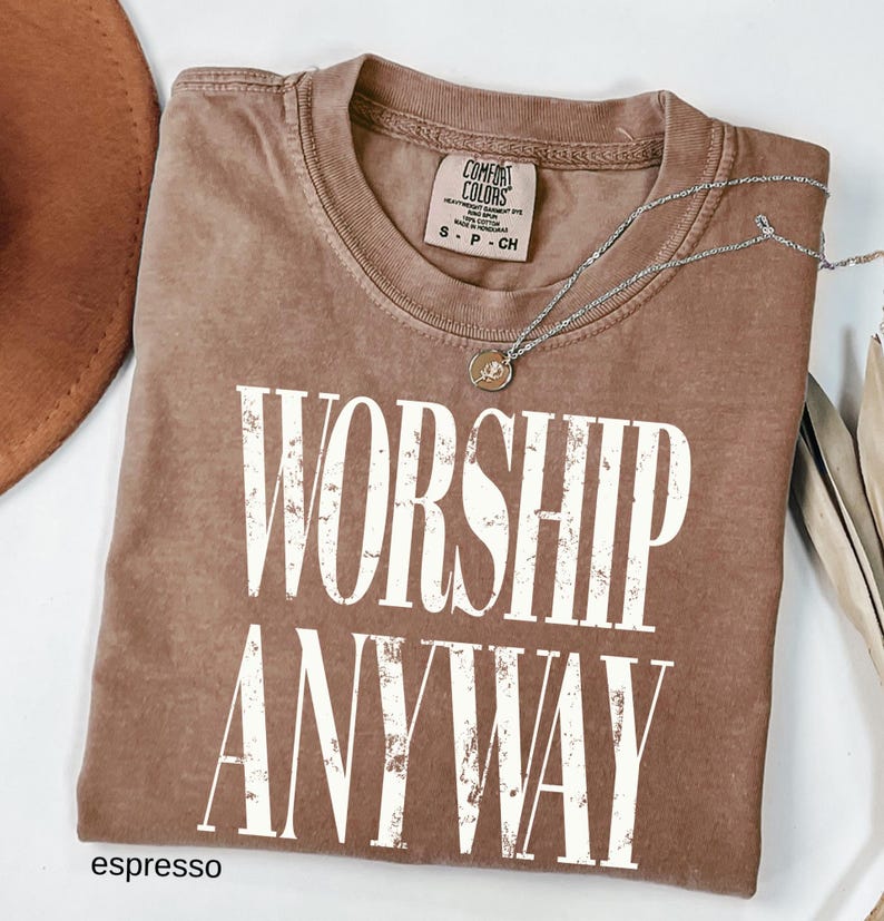 May include: Espresso-colored t-shirt with the words "WORSHIP ANYWAY" in large white letters. The shirt has a crew neck and a small tag at the neckline. A silver necklace with a pendant rests on the shirt. The shirt is folded.