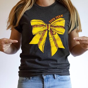 May include: A dark grey t-shirt with a yellow softball bow and the text "YOUR TEAM" printed on the front.
