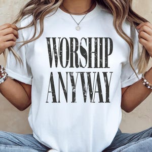 May include: White t-shirt with the text "WORSHIP ANYWAY" in a distressed black font. The shirt is a crew neck style and appears to be made of a soft material. The overall style is casual and comfortable.