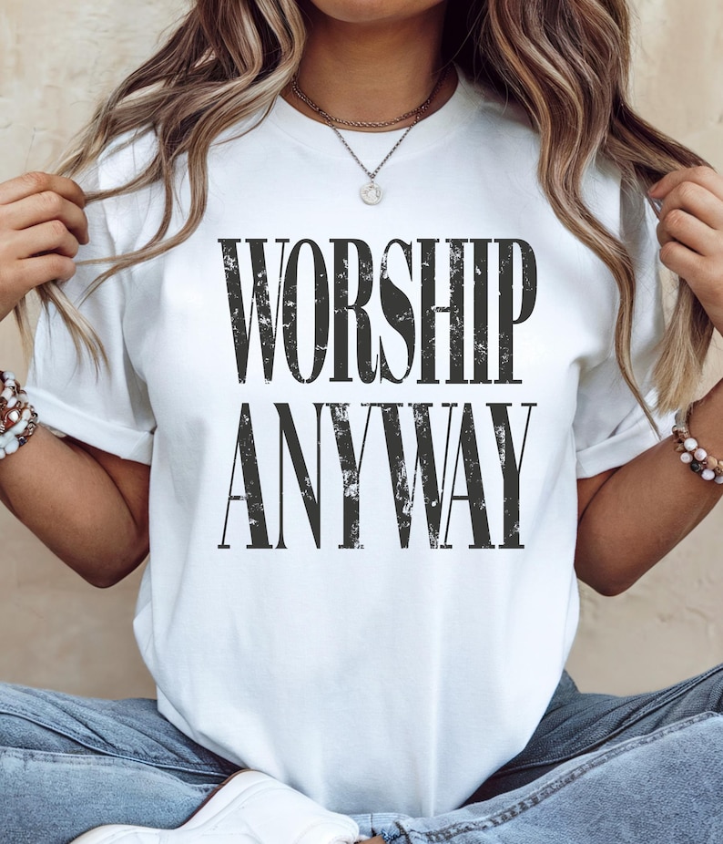 May include: White t-shirt with the text "WORSHIP ANYWAY" in a distressed black font. The shirt is a crew neck style and appears to be made of a soft material. The overall style is casual and comfortable.