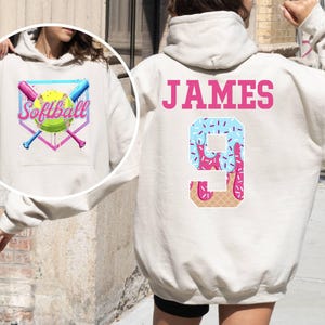 May include: White hoodie with a pink and blue softball design on the front and the text "Softball" and a pink and blue ice cream cone design with the number "9" and the name "JAMES" on the back.