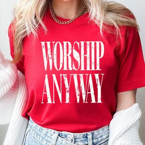 May include: A red t-shirt with the words "WORSHIP ANYWAY" printed in large, distressed white letters. The shirt is short-sleeved and made of a soft material. The model is wearing blue jeans and a gold chain.