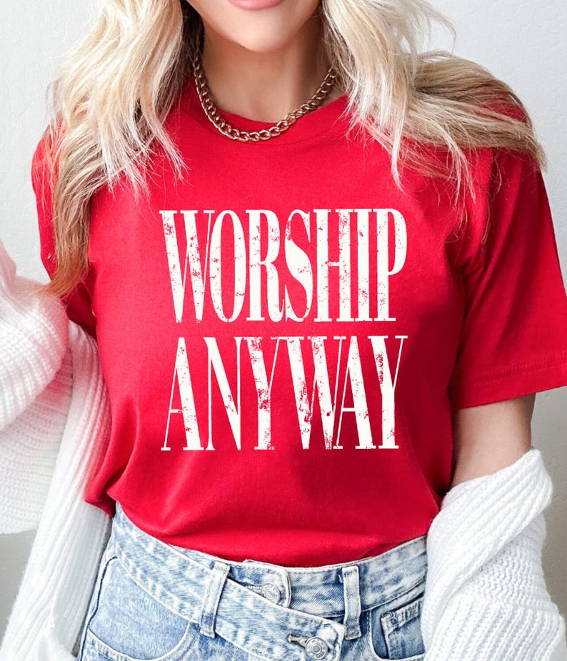 May include: A red t-shirt with the words "WORSHIP ANYWAY" printed in large, distressed white letters. The shirt is short-sleeved and made of a soft material. The model is wearing blue jeans and a gold chain.