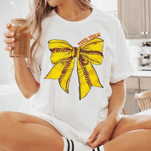 May include: White t-shirt with a yellow softball bow and the text "Your Team" printed on the front.