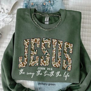 May include: Military green sweatshirt with the word "JESUS" in a camouflage pattern. The text "JOHN 14:6" and "the way the truth the life" are below the word "JESUS". A silver chain is draped on the sweatshirt. The words "military green" are at the bottom.