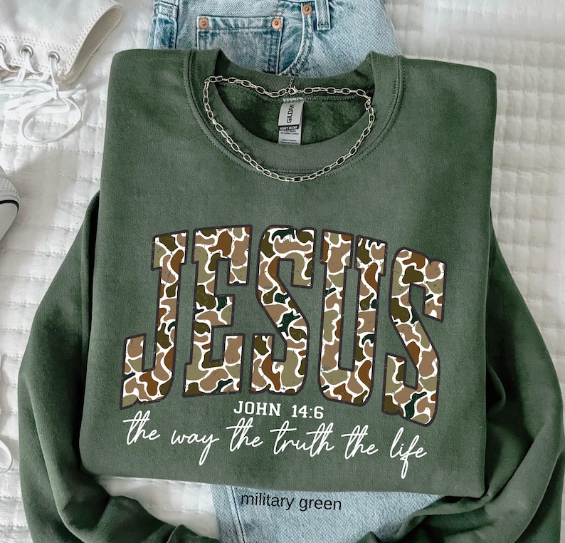May include: Military green sweatshirt with the word "JESUS" in a camouflage pattern. The text "JOHN 14:6" and "the way the truth the life" are below the word "JESUS". A silver chain is draped on the sweatshirt. The words "military green" are at the bottom.