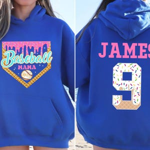 K&ouml;nnte beinhalten: Ein k&ouml;nigsblauer Hoodie mit einem pinkfarbenen und wei&szlig;en Baseball-Motiv auf der Vorderseite. Das Design beinhaltet einen Baseball-Diamanten, einen Baseball und den Text "Baseball Mama". Die R&uuml;ckseite des Hoodies hat ein pinkfarbenes "JAMES" und eine wei&szlig;e Nummer "9" mit bunten Streuseln.