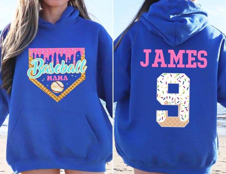K&ouml;nnte beinhalten: Ein k&ouml;nigsblauer Hoodie mit einem pinkfarbenen und wei&szlig;en Baseball-Motiv auf der Vorderseite. Das Design beinhaltet einen Baseball-Diamanten, einen Baseball und den Text "Baseball Mama". Die R&uuml;ckseite des Hoodies hat ein pinkfarbenes "JAMES" und eine wei&szlig;e Nummer "9" mit bunten Streuseln.
