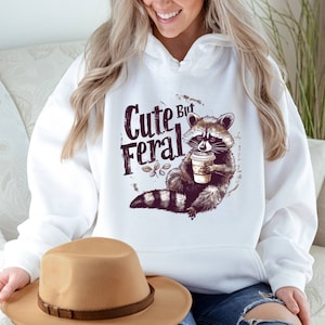 May include: White hoodie with a raccoon graphic and the text "Cute But Feral". The raccoon is holding a cup of coffee.