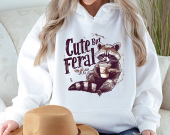 May include: White hoodie with a raccoon graphic and the text "Cute But Feral". The raccoon is holding a cup of coffee.