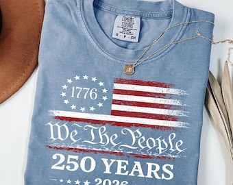 May include: A blue jean colored t-shirt with a distressed American flag design. The shirt features the text "1776", "We The People", "250 YEARS", and "2026". A gold necklace with a pendant is draped on the shirt.
