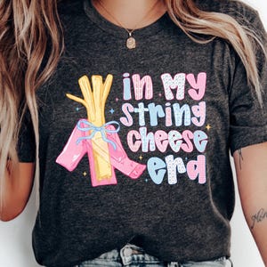 May include: Dark gray t-shirt with the text "in my string cheese era" in colorful letters. The shirt features a graphic of string cheese wrapped in a pink package and tied with a blue ribbon.
