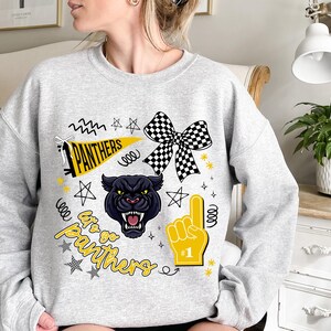 May include: A light gray sweatshirt featuring a black panther graphic with the text "Let's go Panthers." The design includes a checkered bow, a foam finger, stars, and a pennant. The sweatshirt is a comfortable, casual style.