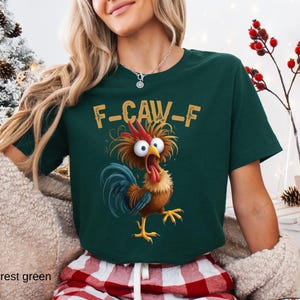 May include: Forest green t-shirt with a cartoon rooster graphic. The rooster has wide eyes, a red comb, and a blue tail. The text "F-CAW-F" is above the rooster. The shirt is paired with red and white plaid pajama pants.