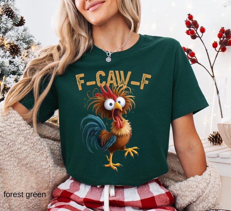 May include: Forest green t-shirt with a cartoon rooster graphic. The rooster has wide eyes, a red comb, and a blue tail. The text "F-CAW-F" is above the rooster. The shirt is paired with red and white plaid pajama pants.