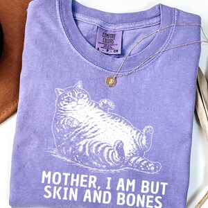 以下が含まれることがあります： 白い猫のイラストと「MOTHER, I AM BUT SKIN AND BONES」の文字が入った紫色のComfort Colors Tシャツ。 金色のペンダント付きネックレスがシャツの上にあります。 シャツは白い表面に折りたたまれています。