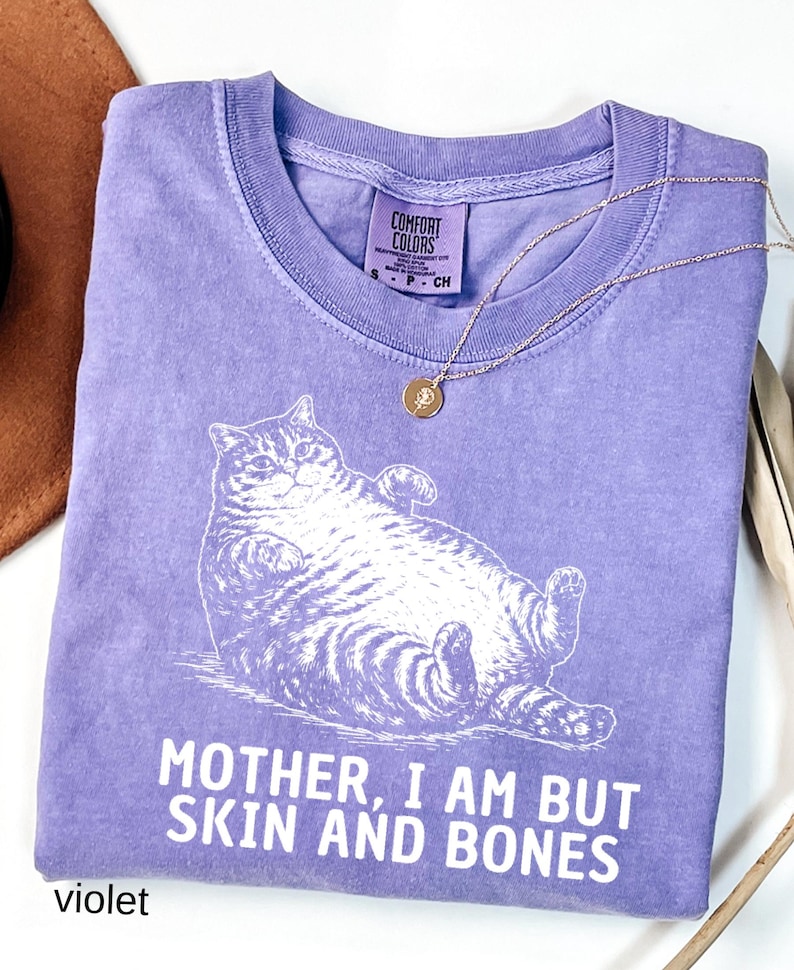 以下が含まれることがあります： 白い猫のイラストと「MOTHER, I AM BUT SKIN AND BONES」の文字が入った紫色のComfort Colors Tシャツ。 金色のペンダント付きネックレスがシャツの上にあります。 シャツは白い表面に折りたたまれています。