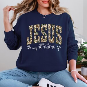 May include: Navy blue sweatshirt with the word "JESUS" in a camouflage pattern. Below the word "JESUS" is the text "JOHN 14:6" and the phrase "the way the truth the life" in a white script font.