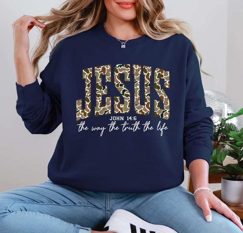 May include: Navy blue sweatshirt with the word "JESUS" in a camouflage pattern. Below the word "JESUS" is the text "JOHN 14:6" and the phrase "the way the truth the life" in a white script font.