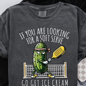 May include: Dark gray t-shirt with a cartoon pickle playing pickleball. The shirt has the text "IF YOU ARE LOOKING FOR A SOFT SERVE GO GET ICE CREAM" in white. The pickle is green with a red headband and yellow shoes.