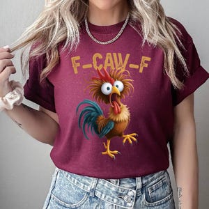 May include: Burgundy t-shirt featuring a cartoon rooster with wide eyes and an open beak. The rooster has blue, brown, and yellow feathers, with the text "F-CAW-F" above it in yellow letters. The shirt is made of a soft material.