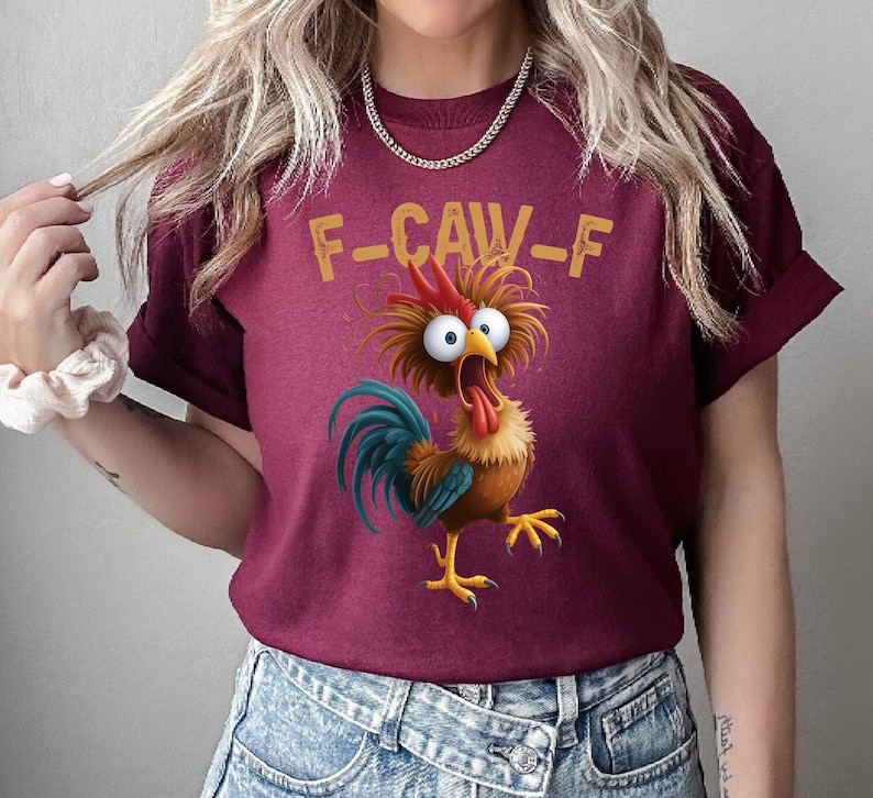 May include: Burgundy t-shirt featuring a cartoon rooster with wide eyes and an open beak. The rooster has blue, brown, and yellow feathers, with the text "F-CAW-F" above it in yellow letters. The shirt is made of a soft material.