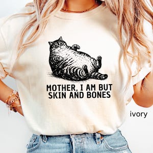 以下が含まれることがあります： アイボリー色のTシャツ。黒と白で描かれた、仰向けに寝ている猫のイラストが特徴です。「MOTHER, I AM BUT SKIN AND BONES」の文字が猫の下にプリントされています。「ivory」の文字が右下にプリントされています。