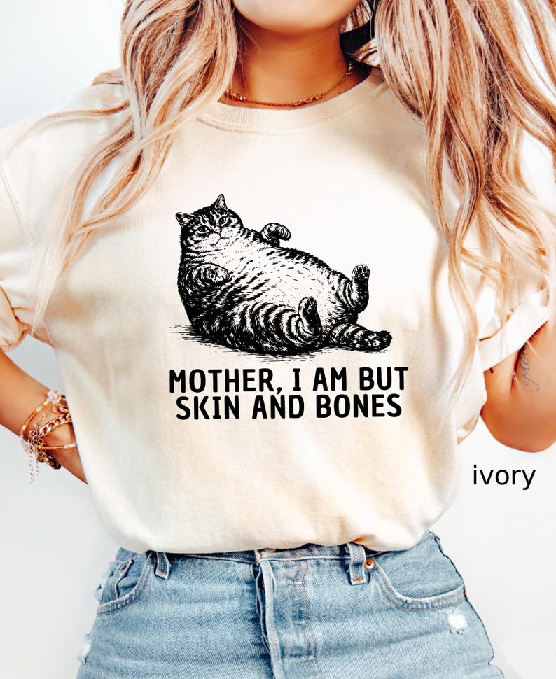 以下が含まれることがあります： アイボリー色のTシャツ。黒と白で描かれた、仰向けに寝ている猫のイラストが特徴です。「MOTHER, I AM BUT SKIN AND BONES」の文字が猫の下にプリントされています。「ivory」の文字が右下にプリントされています。