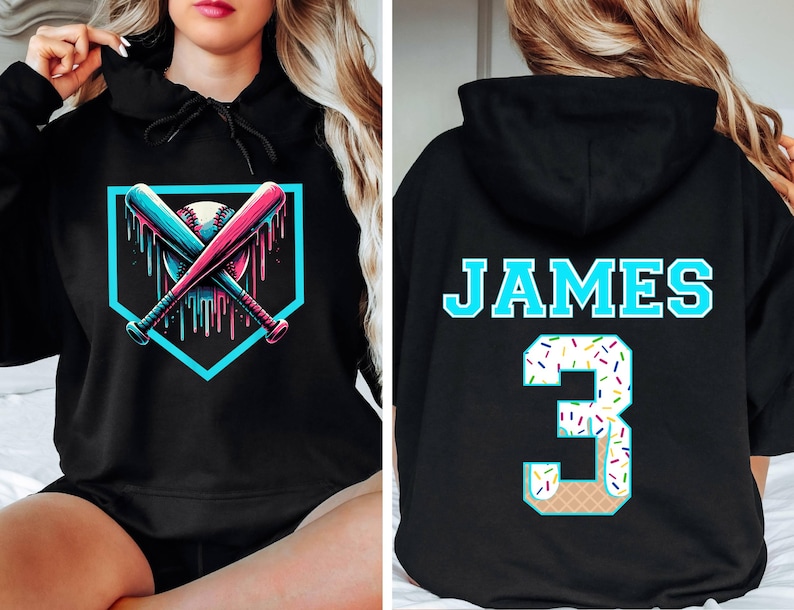 May include: Black hoodie featuring a baseball design with crossed bats and a baseball inside a home plate shape. The bats and dripping accents are blue and pink. The back of the hoodie has the name "JAMES" and the number "3" with a donut-themed design.
