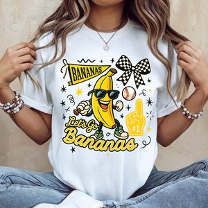 May include: White t-shirt with a cartoon banana wearing sunglasses, holding a baseball bat, and wearing sneakers. The shirt features the text "Let's Go Bananas" and a banner that says "Bananas".
