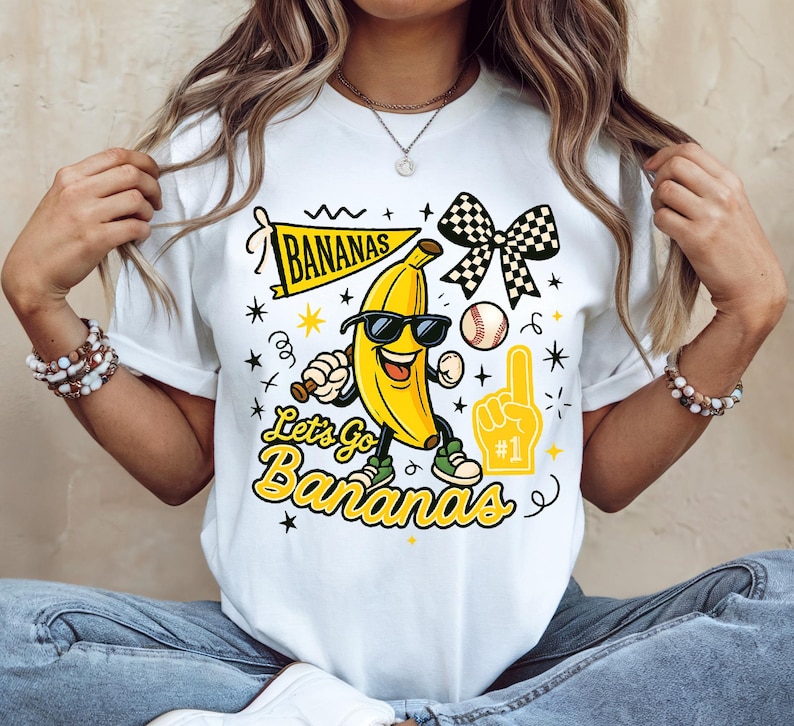 May include: White t-shirt with a cartoon banana wearing sunglasses, holding a baseball bat, and wearing sneakers. The shirt features the text "Let's Go Bananas" and a banner that says "Bananas".