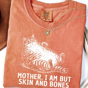 以下が含まれることがあります： リラックスした猫の白いグラフィックと「MOTHER, I AM BUT SKIN AND BONES」というテキストが特徴の、コーラル色のTシャツ。 シャツはクルーネックで、小さなタグが付いています。 ペンダント付きのゴールドネックレスがシャツの上にかけられています。