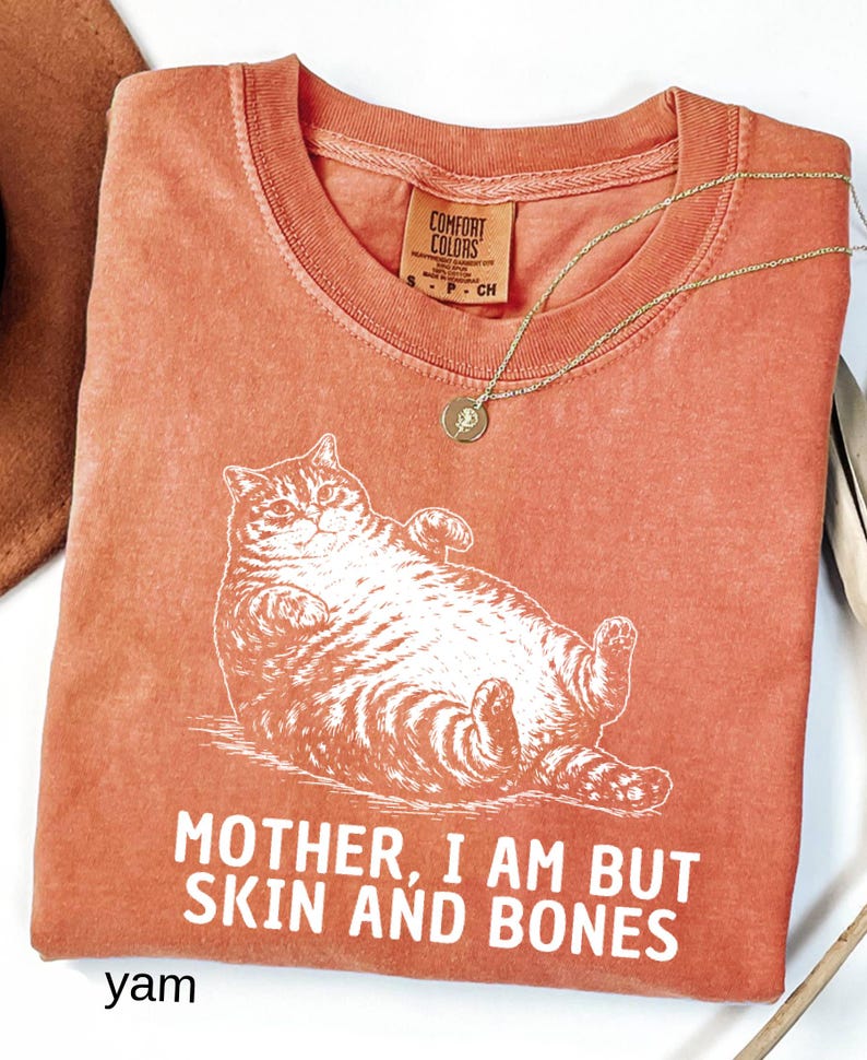 以下が含まれることがあります： リラックスした猫の白いグラフィックと「MOTHER, I AM BUT SKIN AND BONES」というテキストが特徴の、コーラル色のTシャツ。 シャツはクルーネックで、小さなタグが付いています。 ペンダント付きのゴールドネックレスがシャツの上にかけられています。