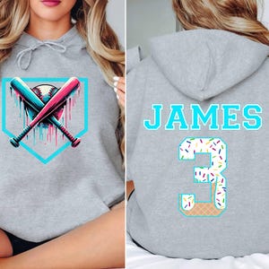 May include: Gray hoodie featuring a baseball design with crossed bats and a baseball, accented with teal and pink. The back displays the name "JAMES" and the number "3" with a waffle cone design.