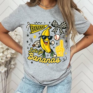 May include: A gray t-shirt featuring a cartoon banana wearing sunglasses and holding a baseball bat. The shirt has the text "Let's Go Bananas" and a banner that says "Bananas". A baseball, foam finger, and checkered bow are also featured.