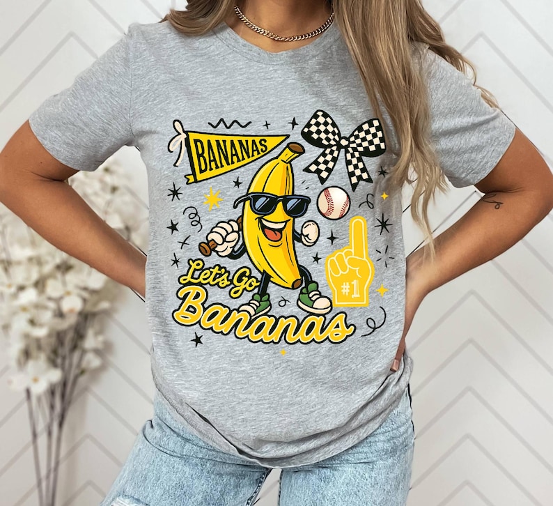 May include: A gray t-shirt featuring a cartoon banana wearing sunglasses and holding a baseball bat. The shirt has the text "Let's Go Bananas" and a banner that says "Bananas". A baseball, foam finger, and checkered bow are also featured.