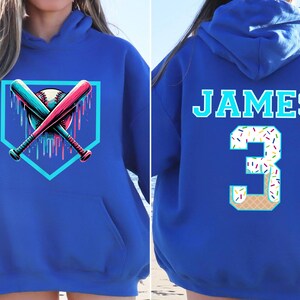 May include: Royal blue hoodie featuring a baseball design on the front with crossed bats and a baseball. The back displays the name "JAMES" and the number "3" designed as an ice cream cone with sprinkles.