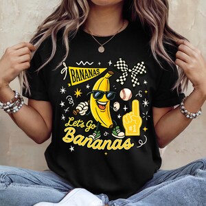 May include: Black t-shirt featuring a smiling banana wearing sunglasses, with the text "Bananas" and "Let's Go Bananas". The design includes a baseball, a foam finger, and a checkered flag. The shirt is a fun, playful design.