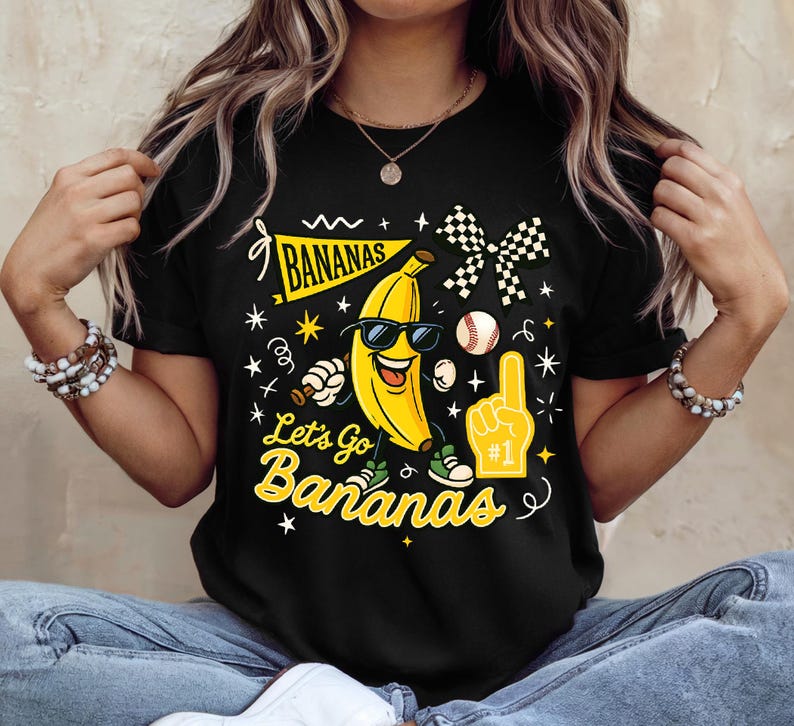 May include: Black t-shirt featuring a smiling banana wearing sunglasses, with the text "Bananas" and "Let's Go Bananas". The design includes a baseball, a foam finger, and a checkered flag. The shirt is a fun, playful design.
