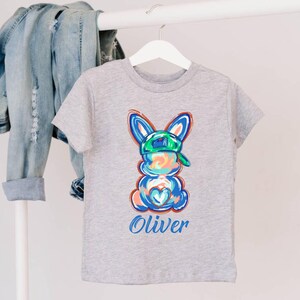 May include: Gray t-shirt with a colorful cartoon bunny wearing a green baseball cap. The bunny has a blue heart on its chest and the name "Oliver" is printed below it.