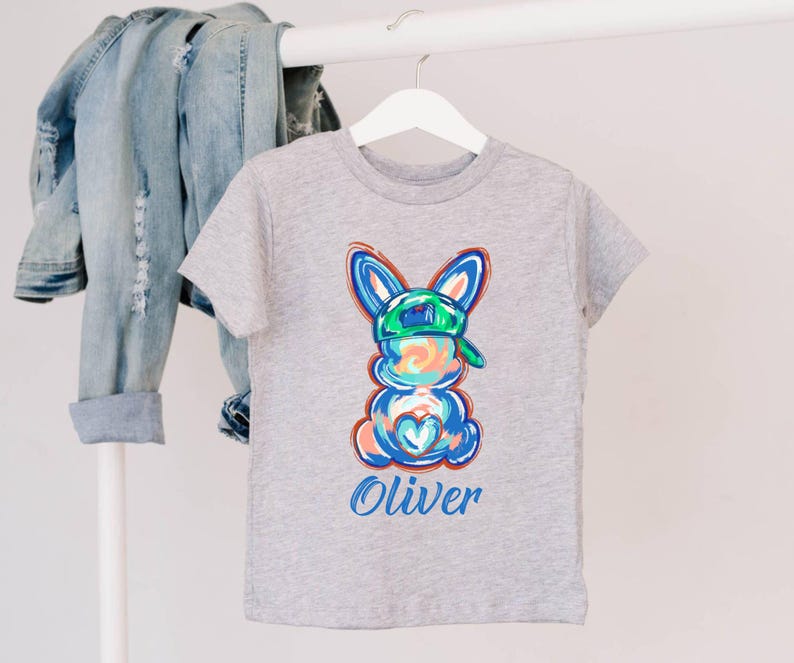 May include: Gray t-shirt with a colorful cartoon bunny wearing a green baseball cap. The bunny has a blue heart on its chest and the name "Oliver" is printed below it.