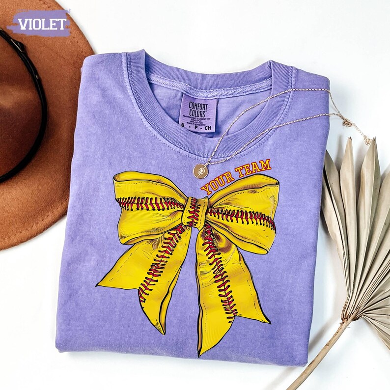 May include: A violet Comfort Colors t-shirt featuring a large yellow softball bow design with red stitching. The text "YOUR TEAM" is printed above the bow. A gold necklace is draped across the shirt.