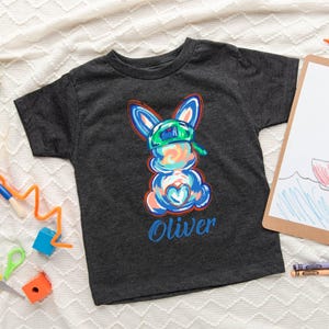 May include: A dark grey t-shirt with a colorful cartoon bunny wearing a green hat. The bunny has a blue heart on its chest and the name "Oliver" is printed below it.